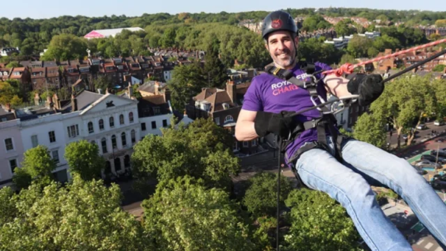 A person in a Royal Free Charity t-shirt and abseil gear, hanging off the side of the Pears Building ready to abseil.