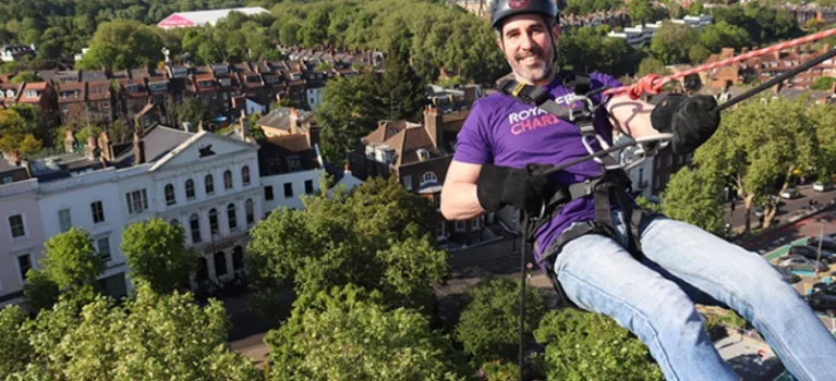 A person in a Royal Free Charity t-shirt and abseil gear, hanging off the side of the Pears Building ready to abseil.